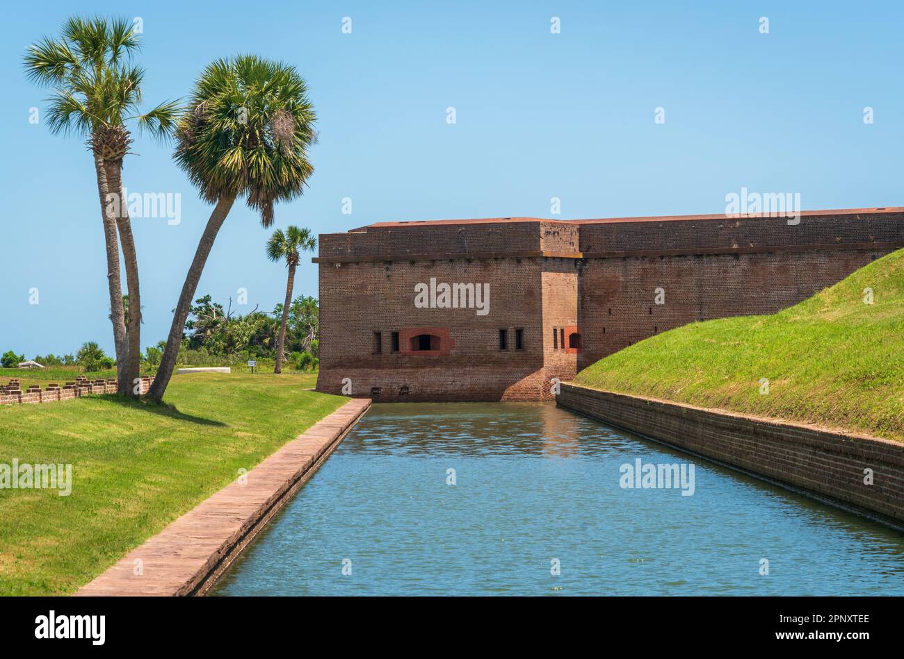 Deux palmiers penchent vers le mur de briques du fort, avec de petites embrasures, au-delà d'un fossé tranquille au Fort Pulaski National Monument, Géorgie. Banque D'Images
