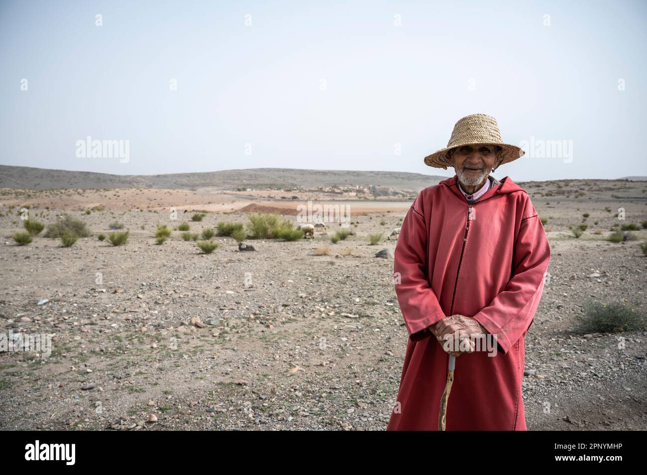 Portrait d'un vieux berger habillé dans un djellaba rouge qui annonce son troupeau dans les terres arides du Maroc. Banque D'Images