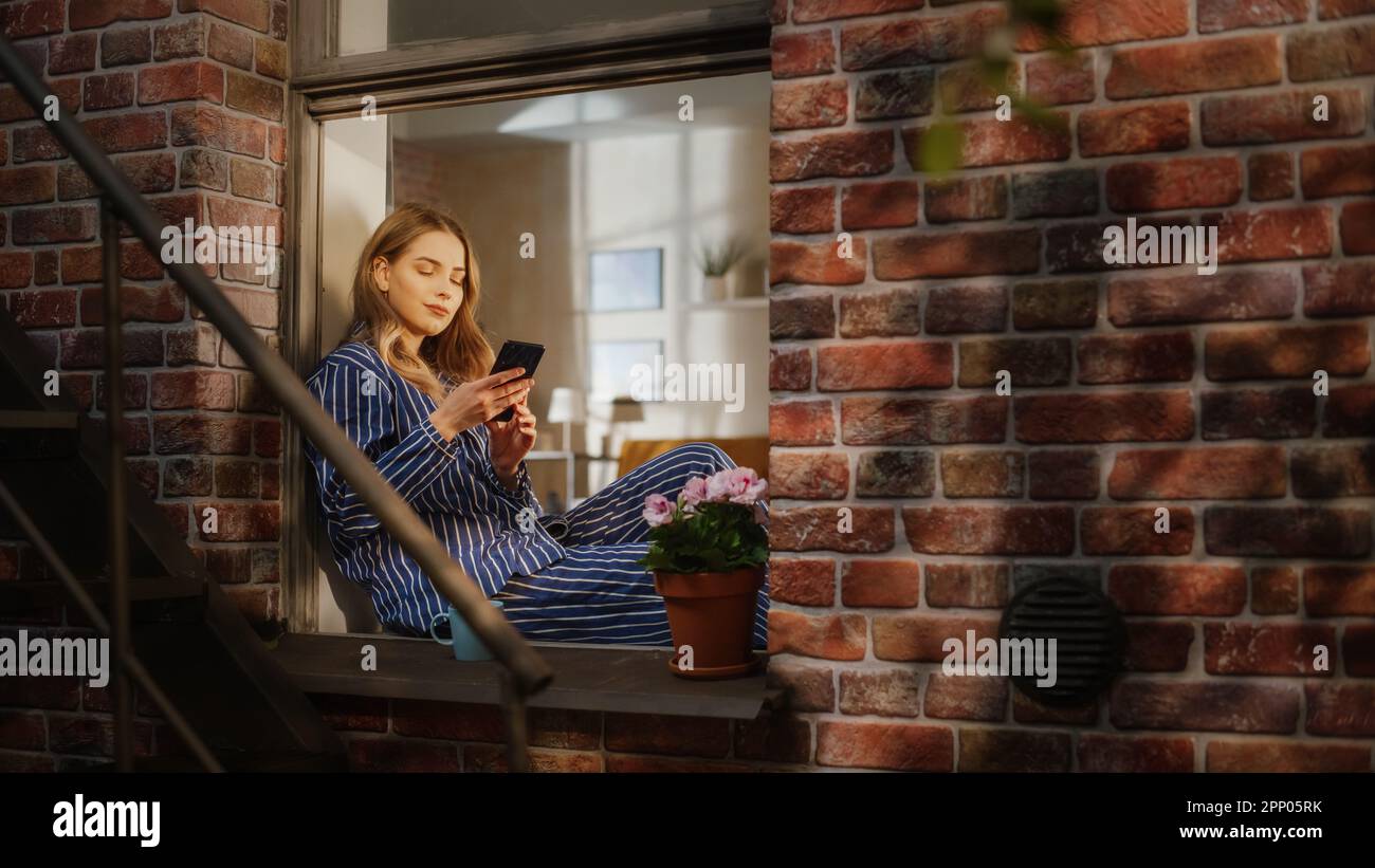 Jeune belle femme utilisant un smartphone et appréciant son café du matin tout en étant assis sur son rebord de fenêtre de la chambre. Pyjama caucasien à rayures pour femme Banque D'Images