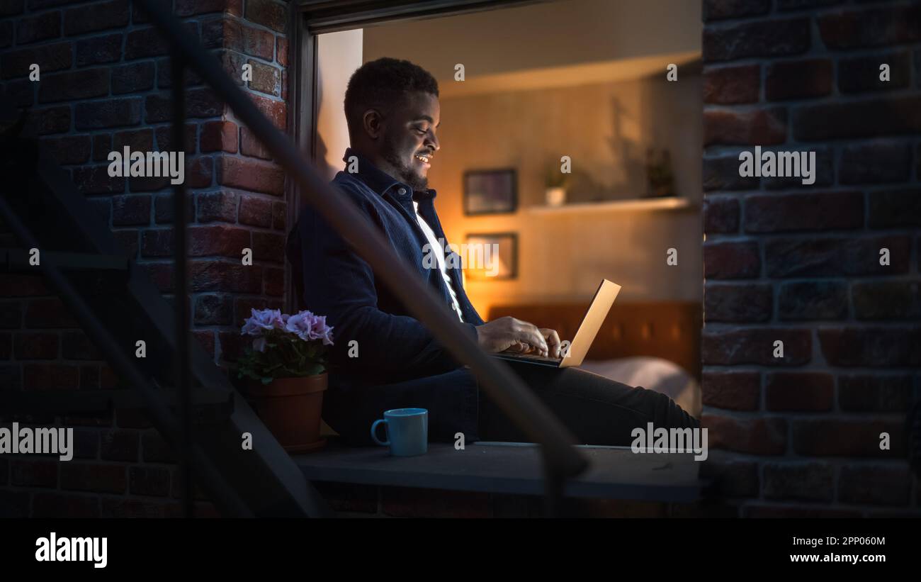 Jeune beau Black Man travaillant à la maison sur ordinateur portable dans la fenêtre d'un confortable appartement loft élégant. Homme souriant, vérifier les médias sociaux Banque D'Images