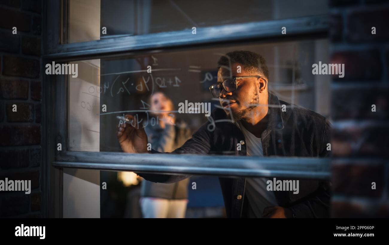 Jeune homme noir expliquant une équation mathématique sur une fenêtre à la maison à un ami féminin en utilisant des marqueurs de verre effaçable. Deux étudiants multiethniques Banque D'Images