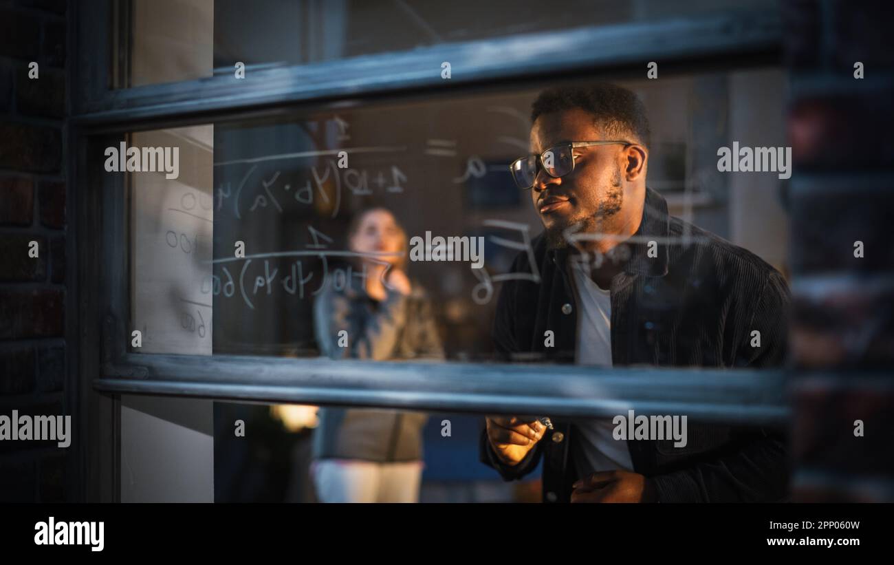 Jeune homme noir expliquant une équation mathématique sur une fenêtre à la maison à un ami féminin en utilisant des marqueurs de verre effaçable. Deux étudiants multiethniques Banque D'Images