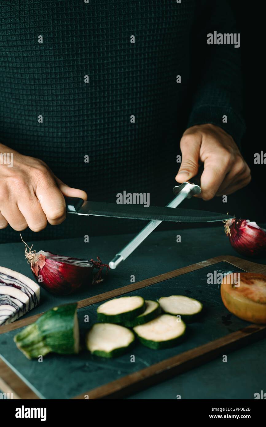 homme à la vapeur d'un couteau de cuisine avec un acier à roder à une table avec quelques légumes hachés Banque D'Images