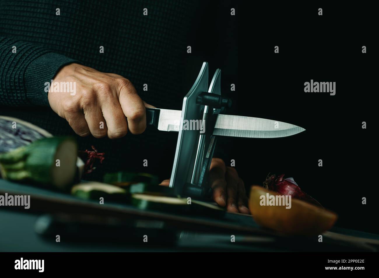 homme aiguisant un couteau de cuisine avec un taille-crayon de bureau sur une table avec un peu de légumes coupés et hachés Banque D'Images