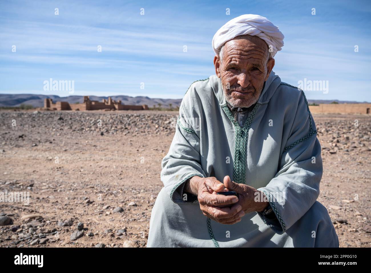 Vieux berbère assis sur une pierre sur le sol du désert à côté des ruines d'une kasbah. Banque D'Images