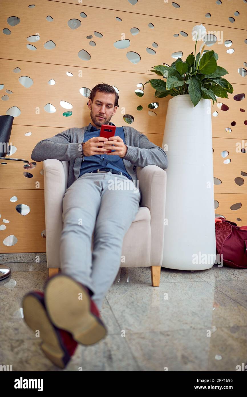 Un jeune homme d'affaires est assis dans le hall de l'hôtel dans une atmosphère détendue et utilise un smartphone. Affaires, personnes, hôtel, voyage Banque D'Images