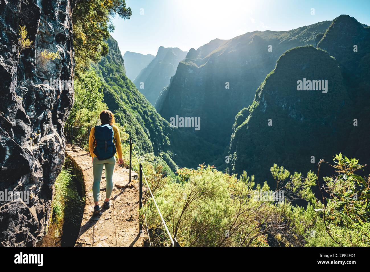 Description: Backpacker femme marchant le long du sentier de randonnée ensoleillé sous le grand mur de roche le long du canal d'eau à la falaise abrupte à travers la forêt tropicale de Madère. Banque D'Images