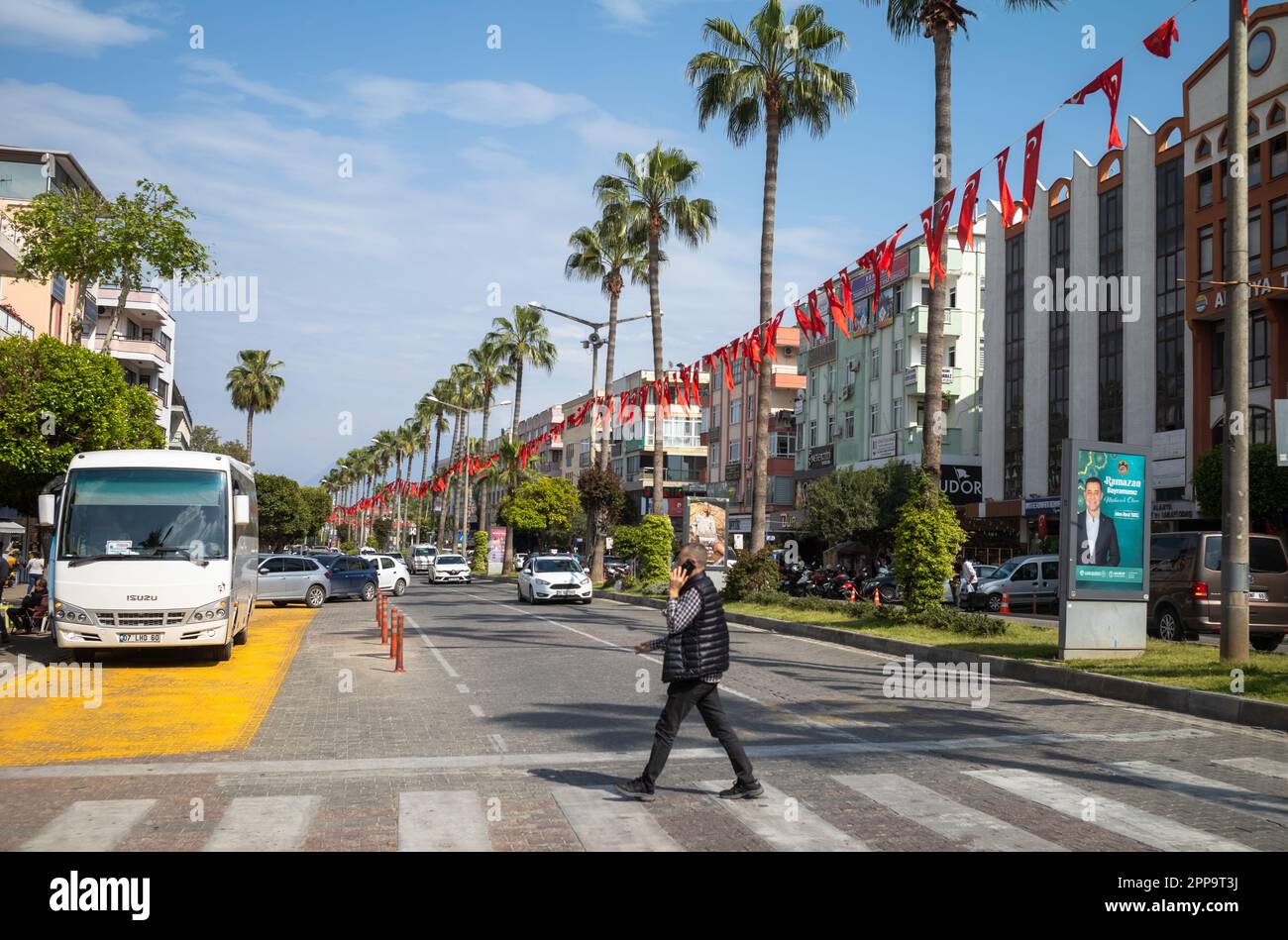 Un homme parlant sur un téléphone portable traverse une route bordée de palmiers et de banderoles avec le drapeau turc à Alanya, dans la province d'Antalya, en Turquie. Banque D'Images