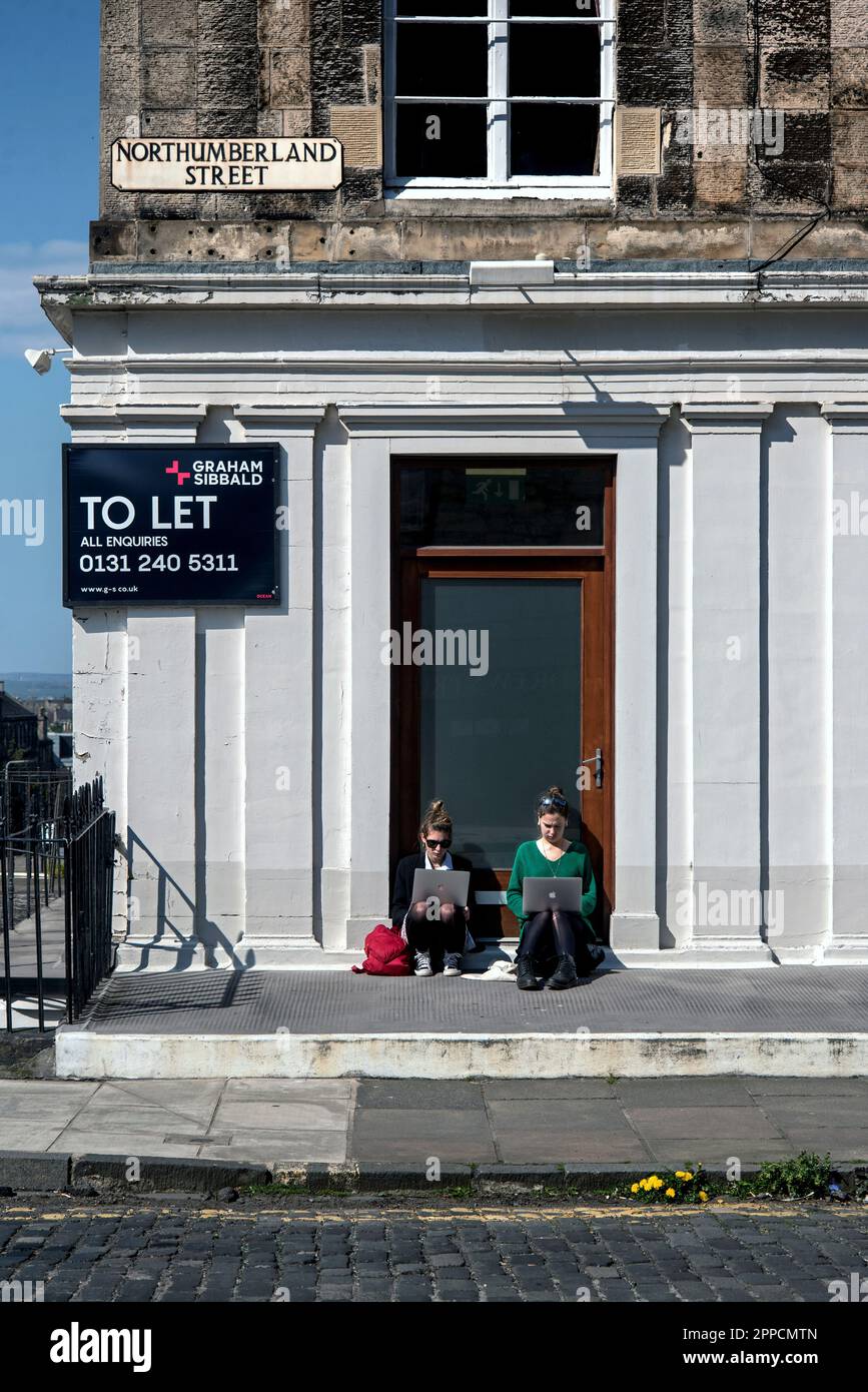 Deux jeunes femmes dans la nouvelle ville d'Édimbourg, assis dans une porte tout en travaillant sur leurs ordinateurs portables et en profitant du soleil de printemps. Banque D'Images