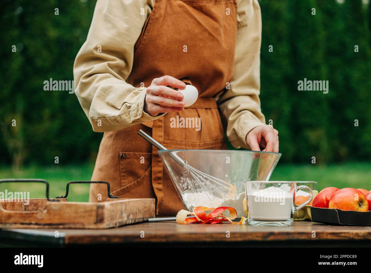Femme en tablier cassant l'œuf dans le bol à mélanger à la table dans le jardin Banque D'Images