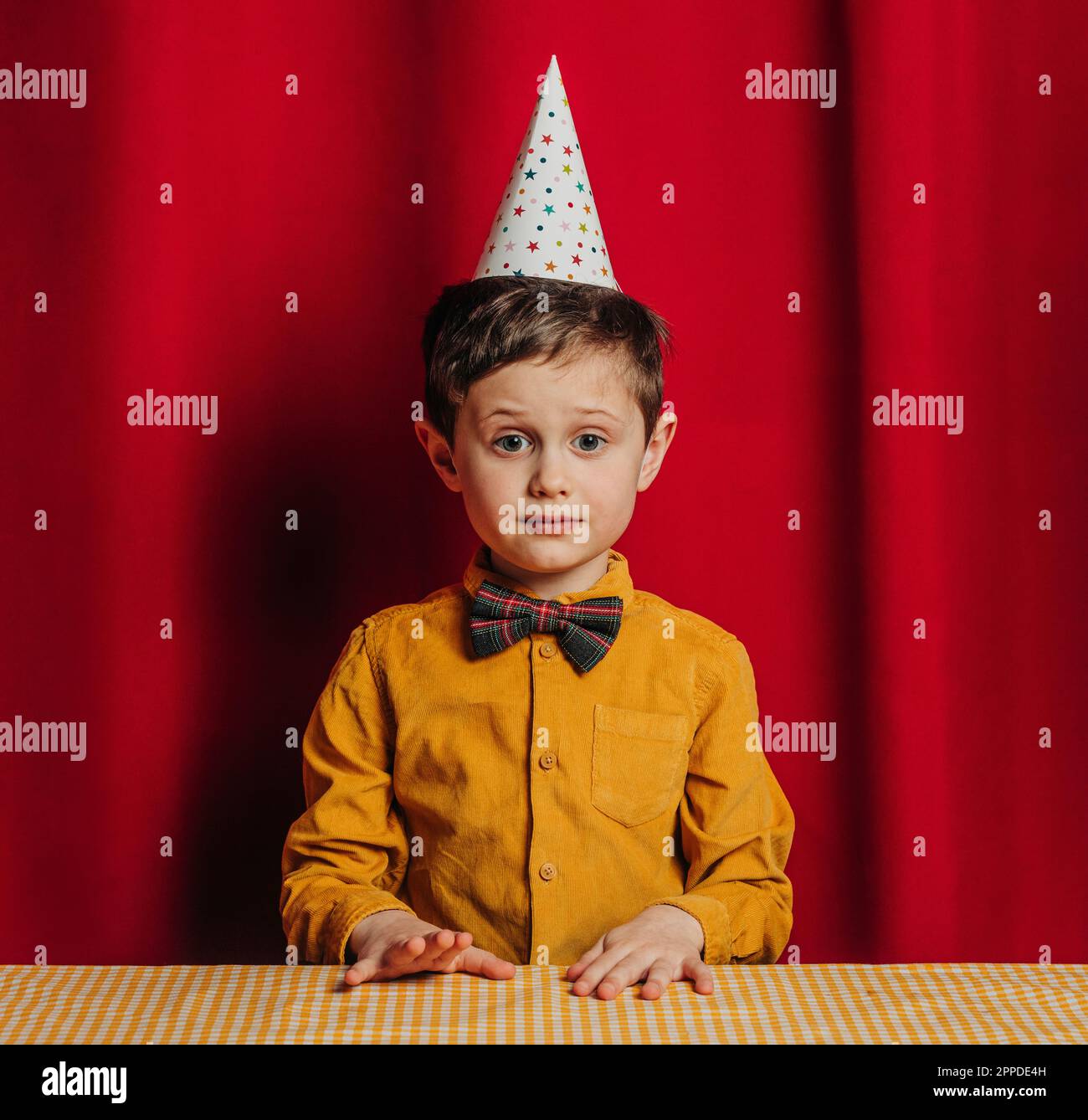 Garçon dans un chapeau d'anniversaire assis à une table devant un rideau rouge Banque D'Images