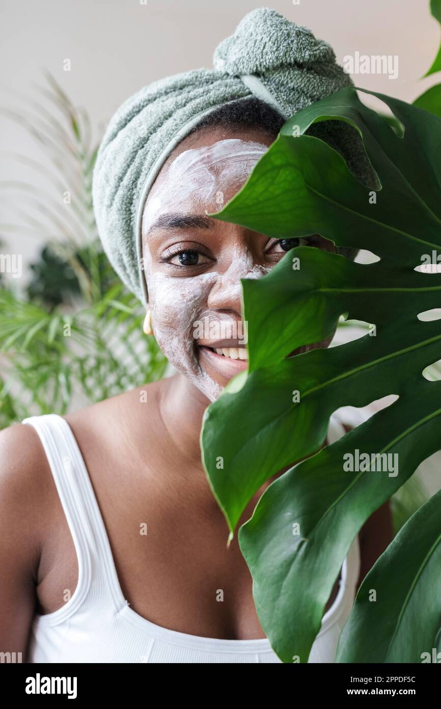 Femme souriante avec masque de visage derrière la feuille de Monstera à la maison Banque D'Images