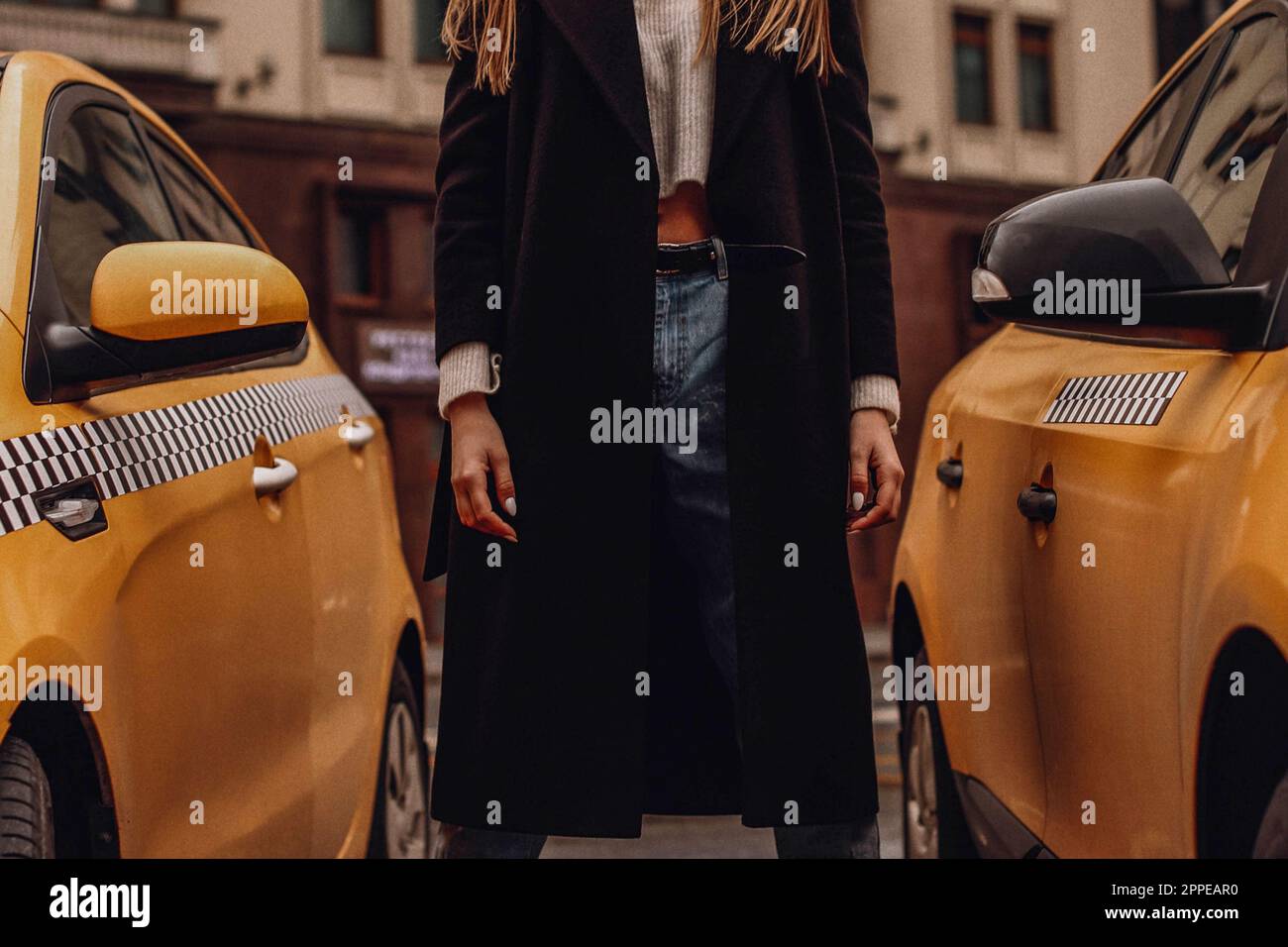 Figure féminine dans un Jean denim bleu et long manteau noir debout contre le taxi jaune. Vêtements d'automne urbains style rue concept Banque D'Images