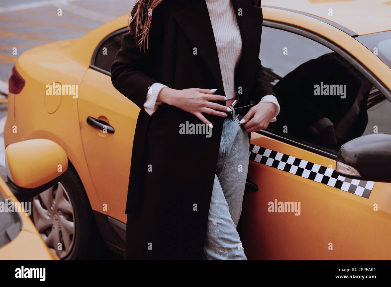 Femme dans un Jean bleu denim, chandail blanc et long manteau noir marchant contre le taxi jaune. Vêtements d'automne urbains style rue concept Banque D'Images