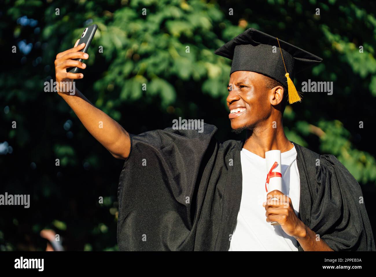Joyeux homme noir diplômé de l'université prenant selfie avec l'appareil photo avant de smartphone avec diplôme d'enseignement supérieur. Étudiant en manteau noir et chapeau Banque D'Images