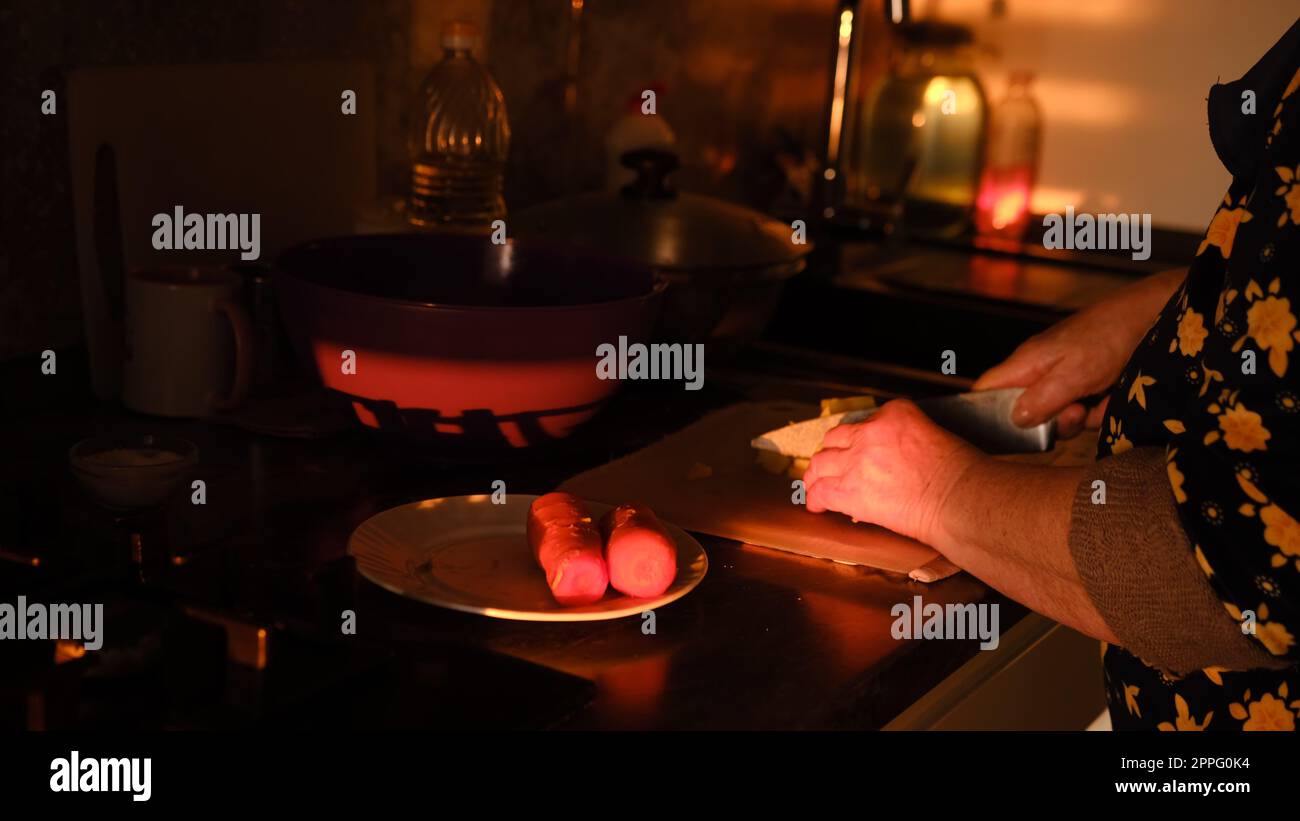 Préparer des légumes sur la cuisine. Les mains de femme senior coupant la carotte bouillie sur la plaque de plastick dans les lumières de coucher de soleil. Vieille femme cuisinant la salade Banque D'Images