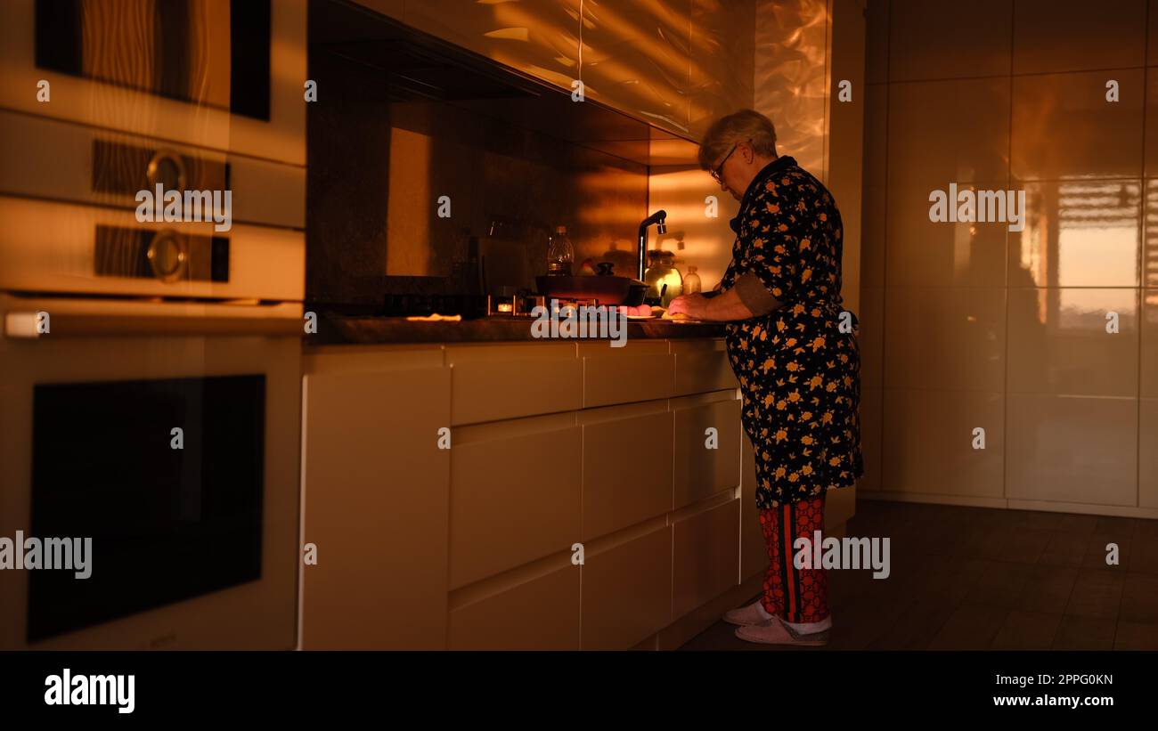 Préparer des légumes sur la cuisine moderne. Femme senior coupant la carotte bouillie sur la plaque de plastick dans les lumières du coucher du soleil. Vieille femme cuisinant la salade Banque D'Images