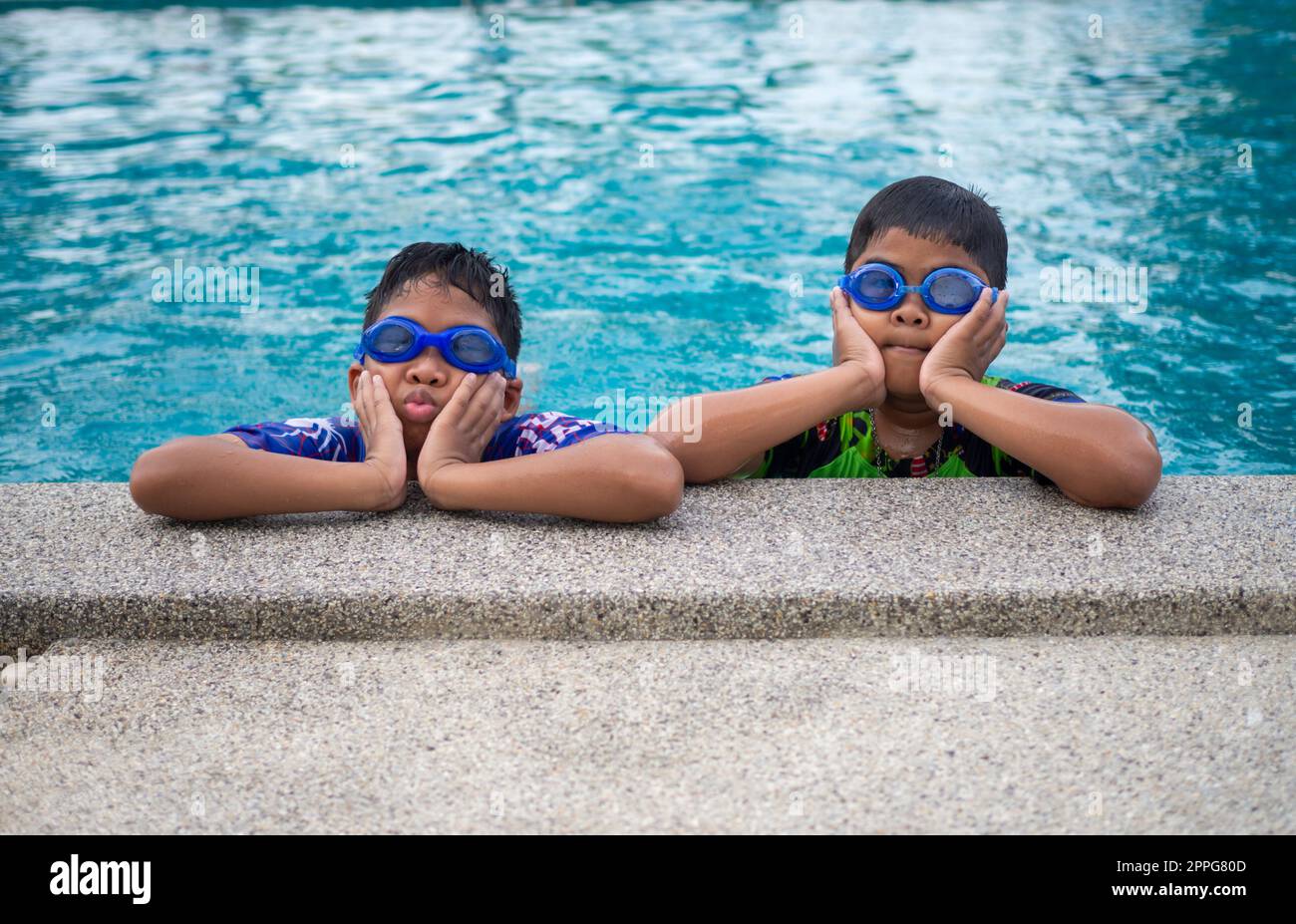 Les frères portant des maillots de bain et des lunettes se sourient tout en étant perchés au bord de la piscine. Banque D'Images