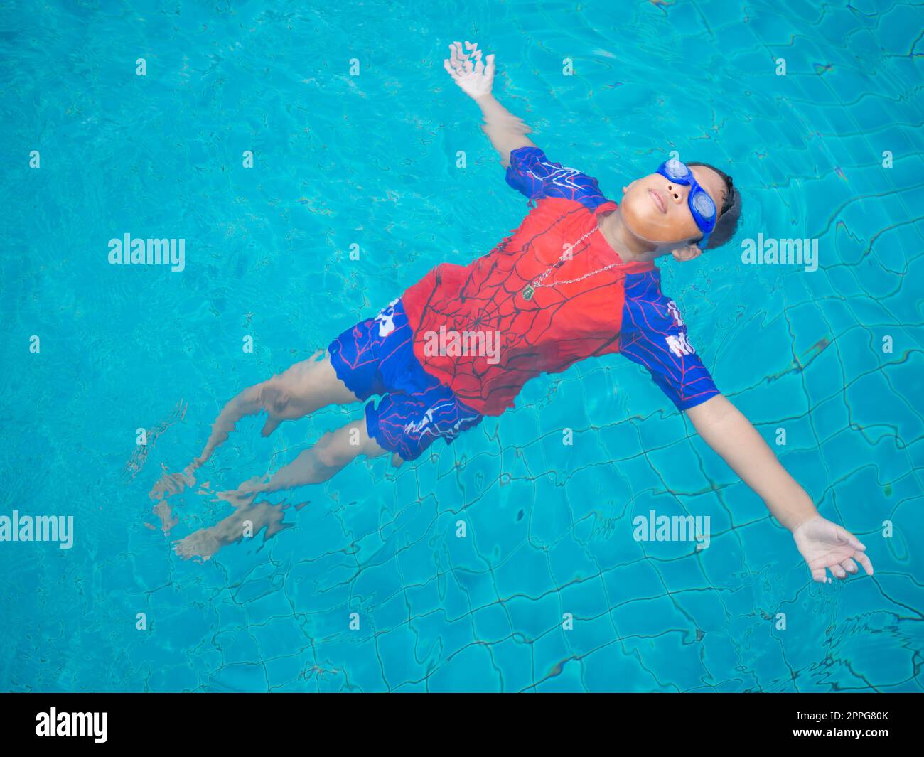 garçon portant un maillot de bain et des lunettes nageant au milieu de la piscine avec un fond bleu Banque D'Images
