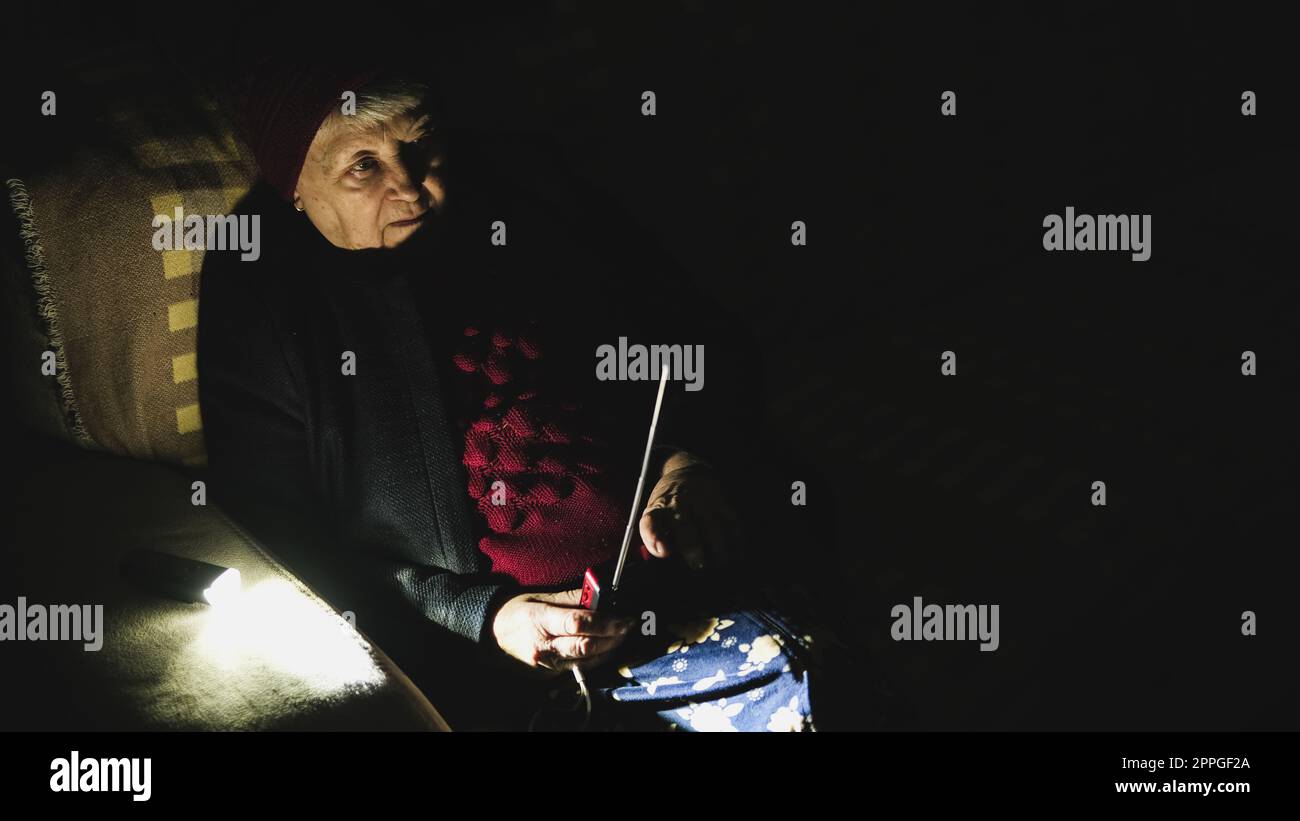 Ukraine : Grand-mère tient la radio dans les mains assis dans l'obscurité blackout pendant les attaques terroristes russes. Femme âgée dans les vêtements d'hiver dans l'obscurité sur le canapé avec récepteur radio à main écoutant des nouvelles Banque D'Images