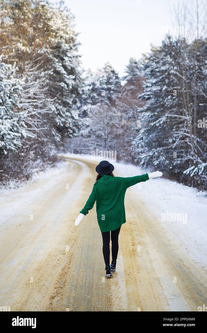 Une jeune fille en chandail vert et chapeau marche au milieu d'une route enneigée dans une épaisse forêt de pins. Journée de congélation. Banque D'Images