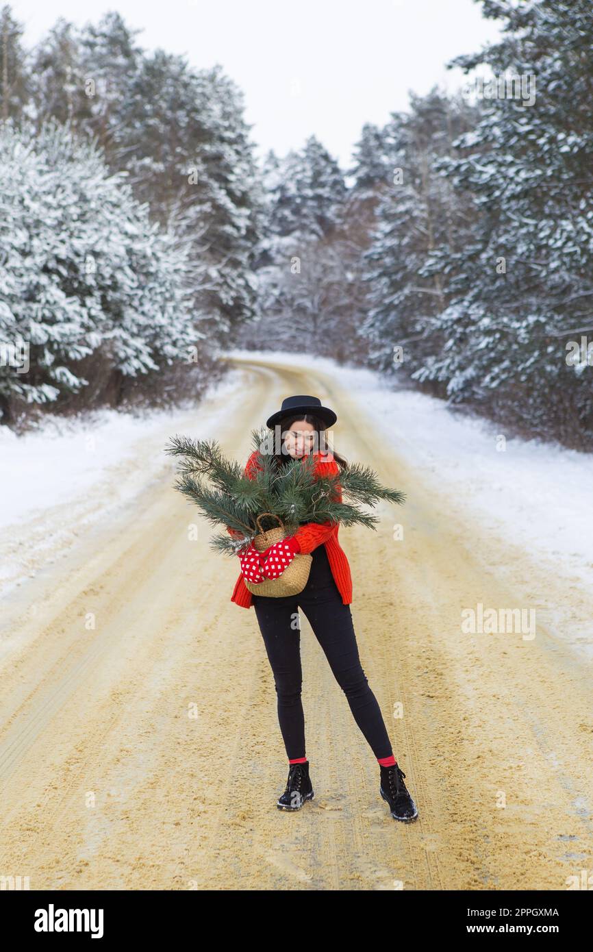 Une fille dans un chandail rouge et un chapeau, en mitaines rouges tient un panier avec des branches de pin dans ses mains se tient au milieu d'une route enneigée dans une forêt avec des branches de pin. Banque D'Images
