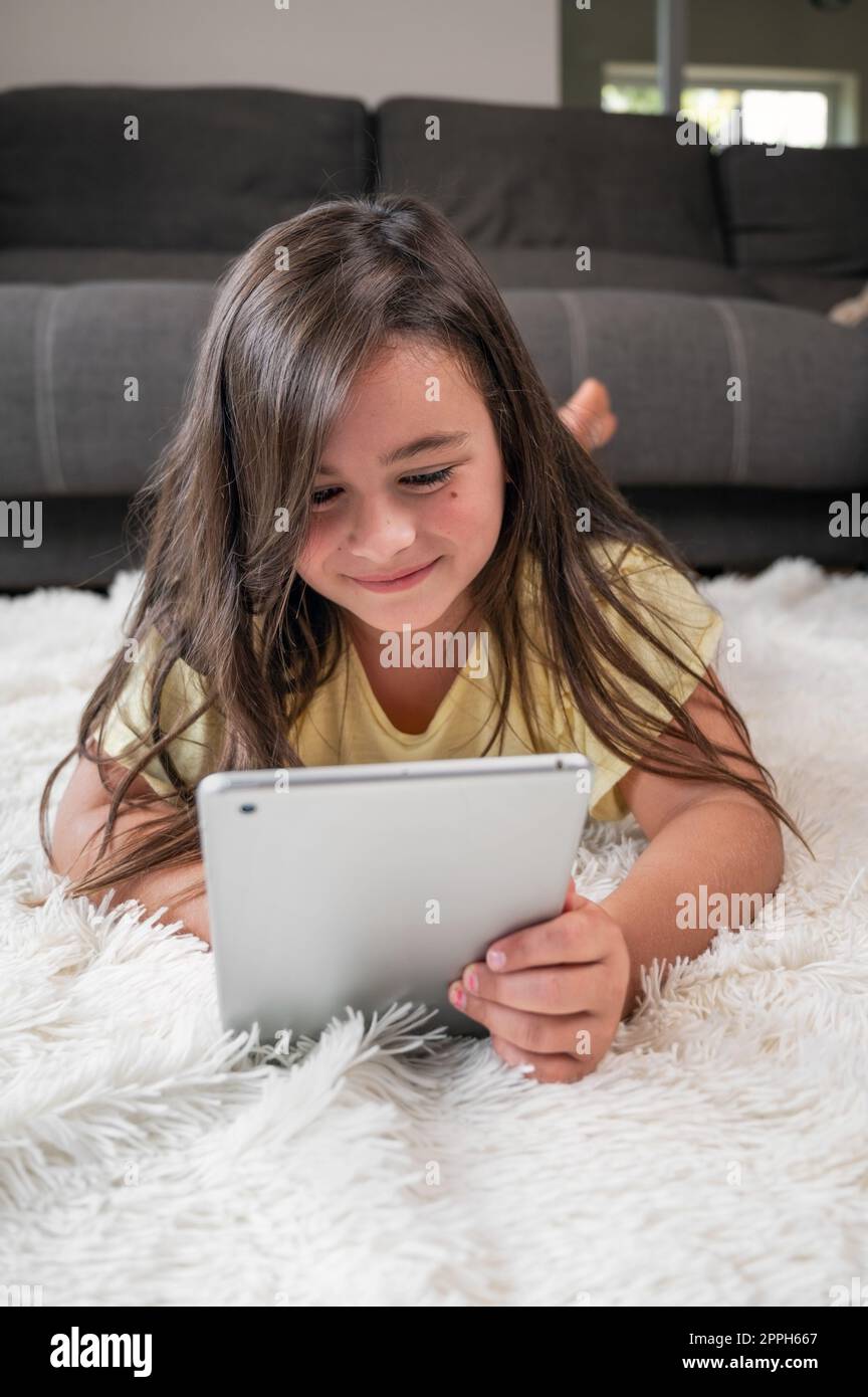 Mignon petite fille regarder des dessins animés sur tablette numérique. Enfant allongé sur le sol riant à l'aide d'un appareil électronique. Loisirs intérieurs pour les enfants. Banque D'Images