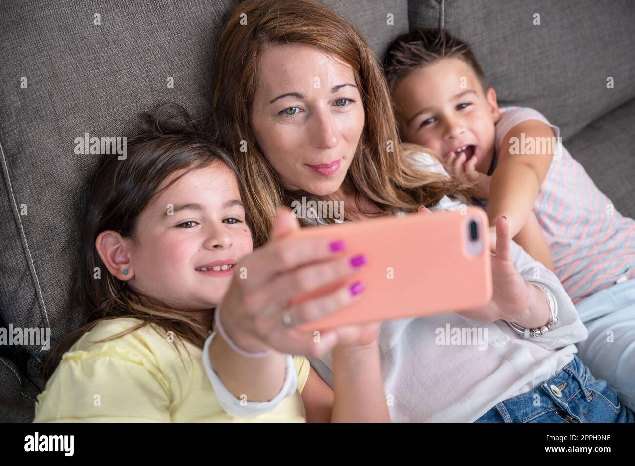 Mère heureuse avec ses enfants font un selfie ou un appel vidéo au père ou à des parents dans un canapé. Concept de technologie, nouvelle génération, famille, connexion, parentalité. Banque D'Images