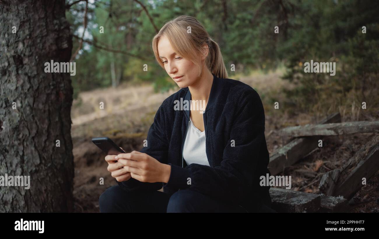 Portrait d'une jeune belle femme Blonde dans une atmosphère romantique nature. Elle est vêtue de noir et utilise son smartphone. Elle naviguant quelque peu Banque D'Images