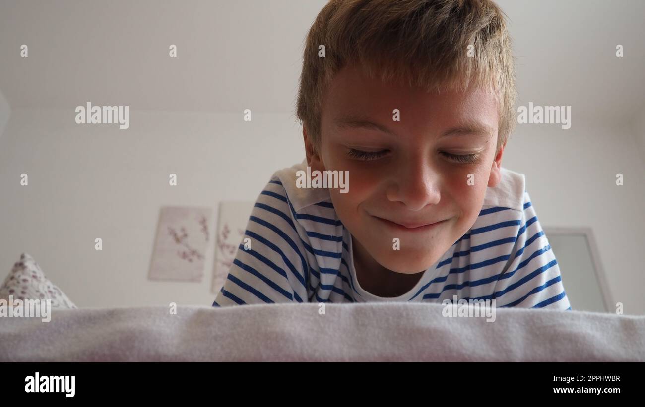 Mignon petit garçon caucasien de 10 ans se trouve à la maison sur le lit dans la chambre et sourit slyly, plissant et fermant les yeux. Attendre une surprise, une blague et se faire dorloter. Peekaboo. Jouez à Blinders. Banque D'Images