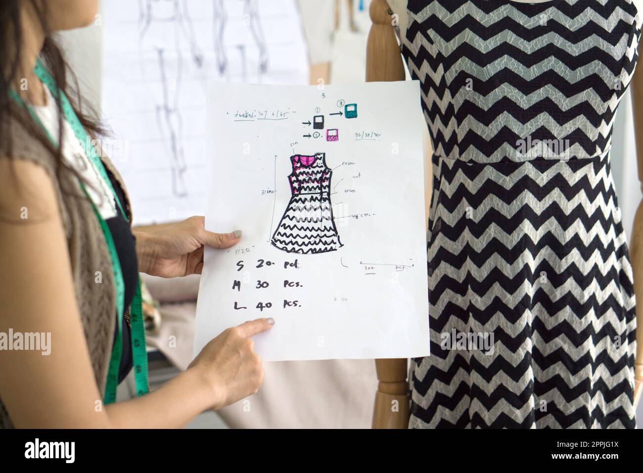 Jeune propriétaire d'entreprise de design de mode vérifie les commandes de la note sur le modèle pour le tissu, en comparant avec la robe sur le mannequin. Banque D'Images