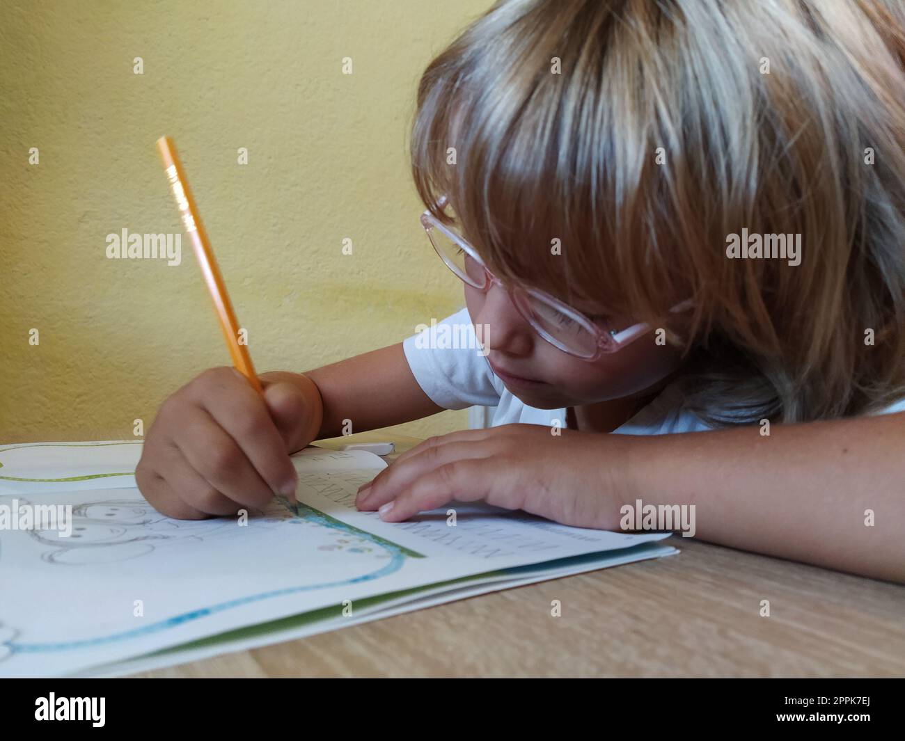 Une fille de 6 ou 7 ans dessine dans un livre. L'enfant fait ses devoirs. Cheveux blonds et lunettes à bordure rose. Concept de formation et d'éducation Banque D'Images