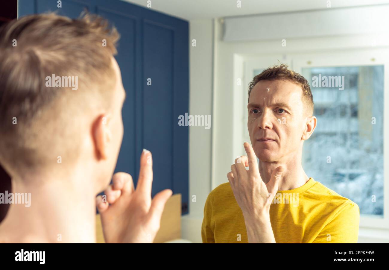 Homme mature appliquer la crème hydratante avec le doigt sur les zones claires du visage. Portrait masculin dans le miroir de la chambre. Concept de soin du visage Banque D'Images