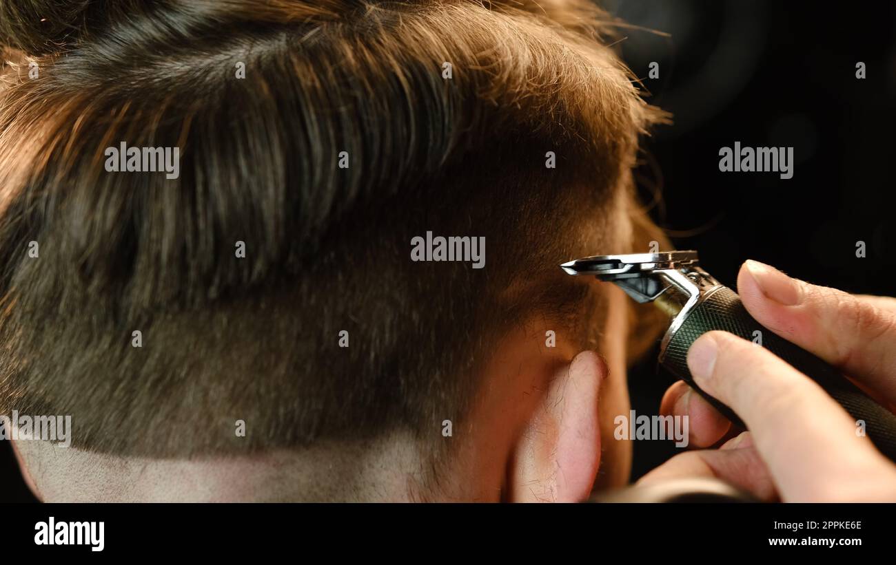 Gros plan photo d'un beau barbier donnant une coupe de cheveux à son client à l'aide d'un coupe-bordure. Salon de coiffure dans un salon de coiffure moderne dans un éclairage sombre avec une lumière chaleureuse et une vue arrière Banque D'Images