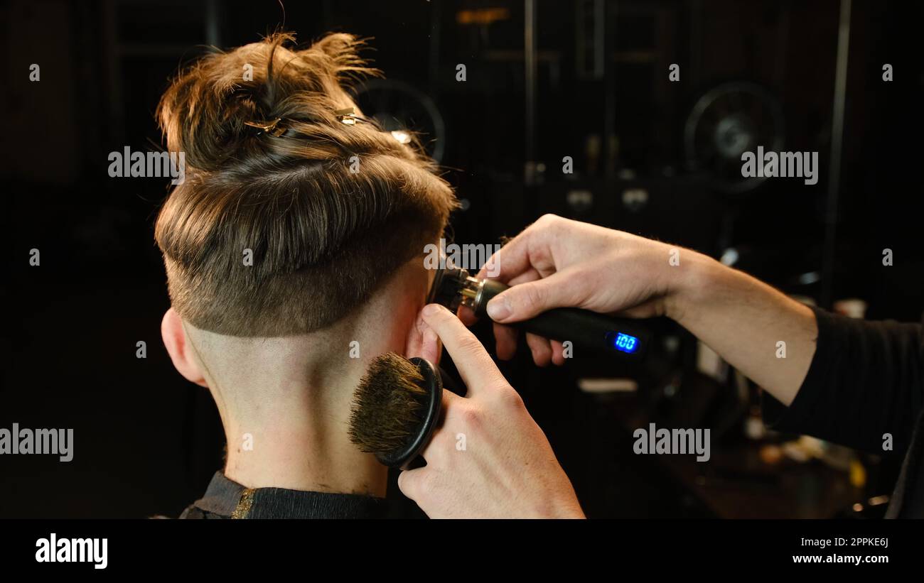 Photo d'un beau coiffeur donnant une coupe de cheveux à son client à l'aide d'une tondeuse. Service de coiffeur dans un salon de coiffure moderne dans un éclairage sombre clé avec vue arrière de lumière chaude Banque D'Images