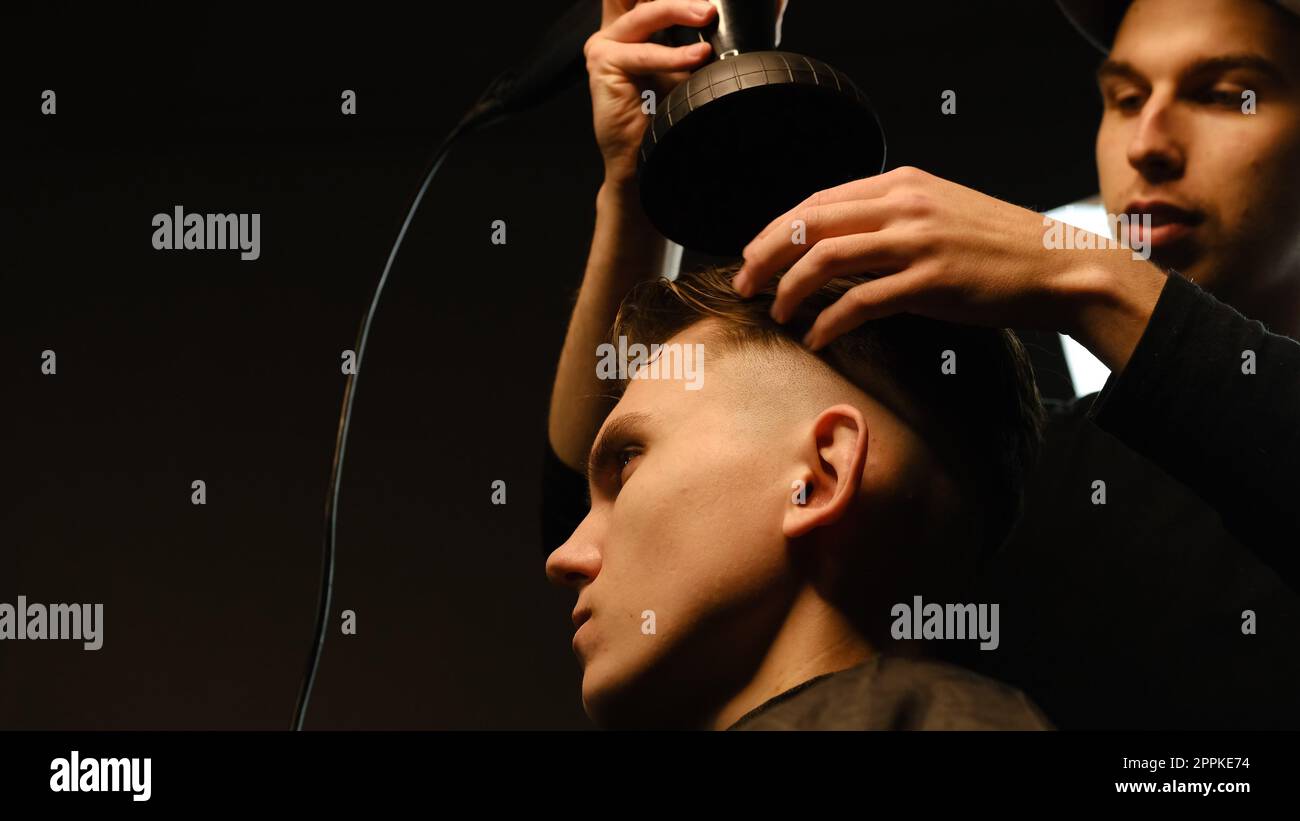 Jeune coiffeur séchant les cheveux du client avec sèche-cheveux dans le salon de coiffure dans un éclairage de clé sombre avec la lumière chaude et les cheveux corrects avec la main. Coiffeur pour homme Banque D'Images