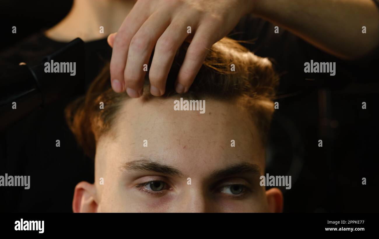 Close up jeune barbier sèche les cheveux du client avec sèche-cheveux dans le salon de coiffure dans un éclairage sombre clé avec la lumière chaude et les cheveux corrects avec la main. Coiffeur pour homme vue de face Banque D'Images
