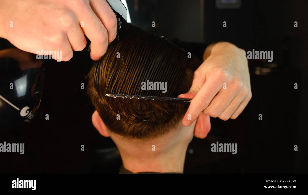 Pulvérisation de cheveux. barbier pulvérisant de l'eau sur les cheveux du client et peignez-le pour la coupe et le coiffage. Concept Barbershop. vue arrière en éclairage sombre clé avec lumière chaude Banque D'Images