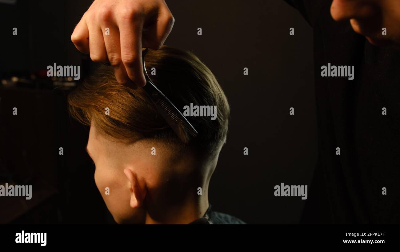 Barber utiliser des ciseaux d'amincissement et peigne en métal sur les cheveux ondulés bruns du jeune homme. Service de coiffeur dans un salon de coiffure moderne dans un éclairage de clé sombre avec une lumière chaude Banque D'Images