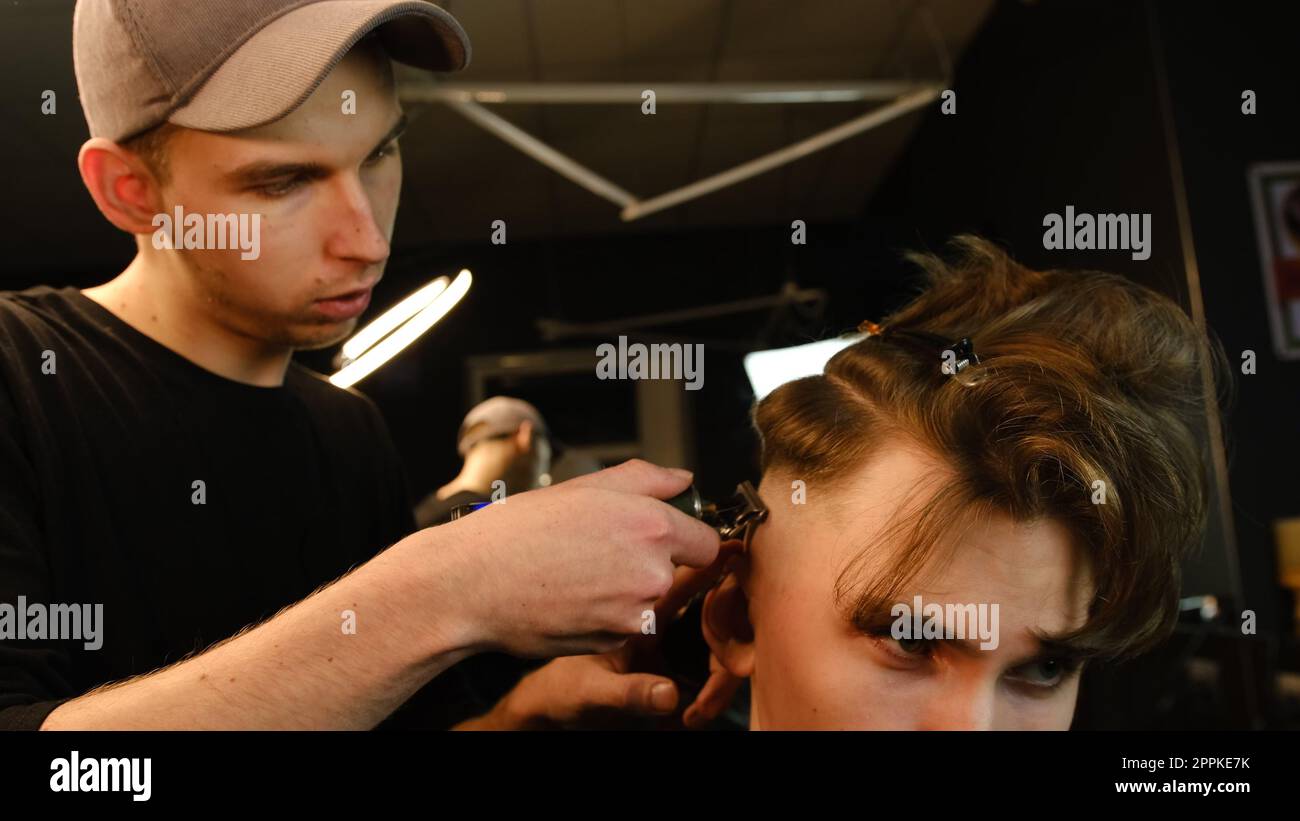 Photo d'un beau coiffeur donnant une coupe de cheveux à son client à l'aide d'une tondeuse. Service de coiffeur dans un salon de coiffure moderne dans un éclairage sombre clé avec vue latérale de lumière chaude Banque D'Images