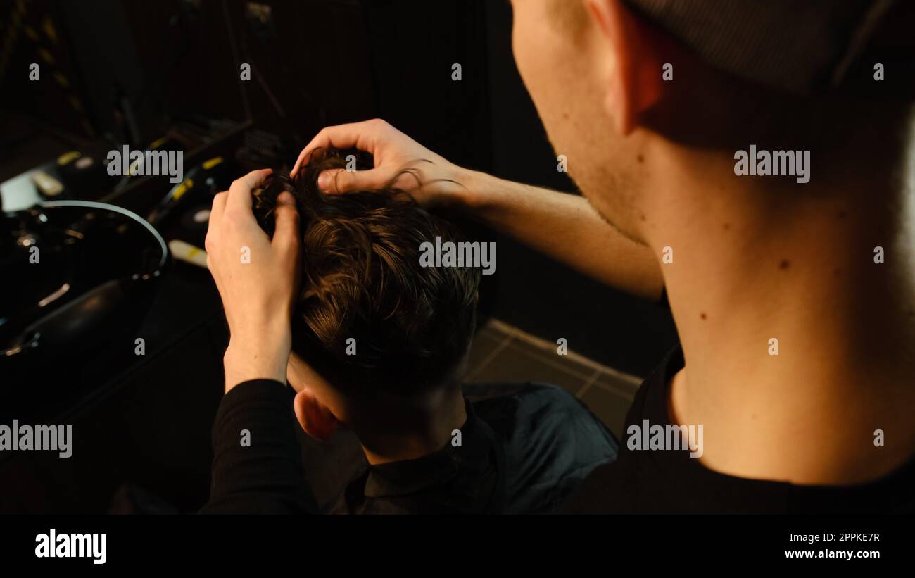 Salon de coiffure. Homme dans la chaise de barbier, coiffeur coiffant ses cheveux avec les mains dans l'éclair sombre clé. Coiffeur pour homme Banque D'Images