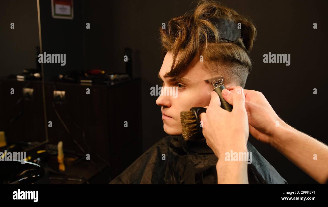 Photo d'un beau coiffeur donnant une coupe de cheveux à son client à l'aide d'une tondeuse. Service de coiffeur dans un salon de coiffure moderne dans un éclairage sombre clé avec vue latérale de lumière chaude Banque D'Images