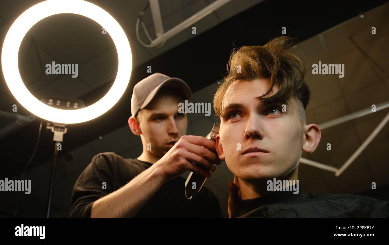 coiffure pour hommes et coupe de cheveux avec tondeuse à cheveux dans un salon de coiffure ou de coiffure. Service de coiffeur dans un salon de coiffure moderne dans un éclairage sombre clé avec lumière chaude vue à faible angle Banque D'Images