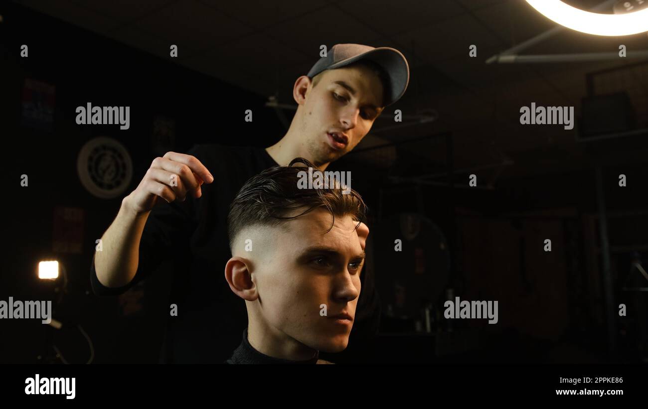 Salon de coiffure. Homme dans la chaise de barbier, coiffeur coiffant ses cheveux avec les mains dans l'éclair sombre clé. Coiffeur pour homme Banque D'Images