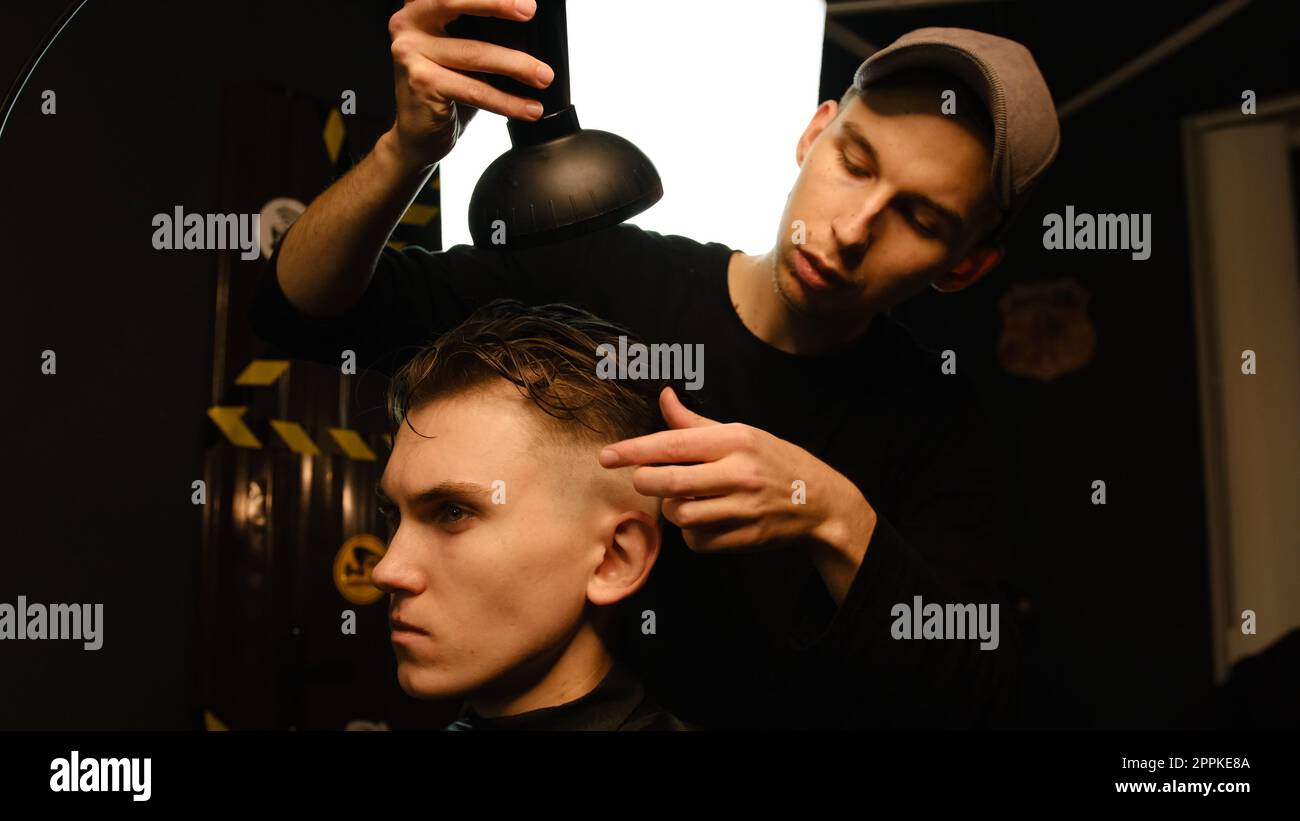 Jeune coiffeur séchant les cheveux du client avec sèche-cheveux dans le salon de coiffure dans un éclairage de clé sombre avec la lumière chaude et les cheveux corrects avec la main. Coiffeur pour homme Banque D'Images