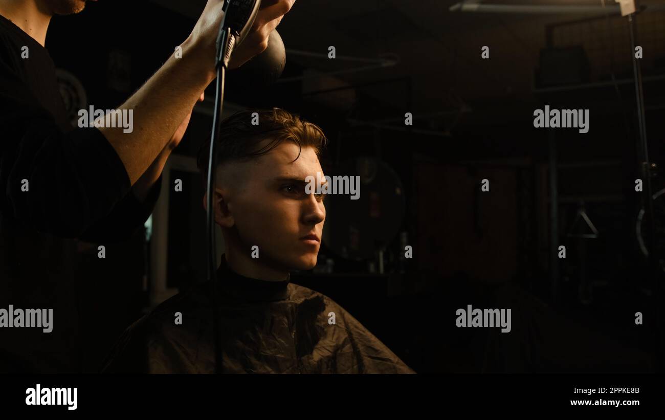 Jeune coiffeur séchant les cheveux du client avec sèche-cheveux dans le salon de coiffure dans un éclairage de clé sombre avec la lumière chaude et les cheveux corrects avec la main. Coiffeur pour homme Banque D'Images