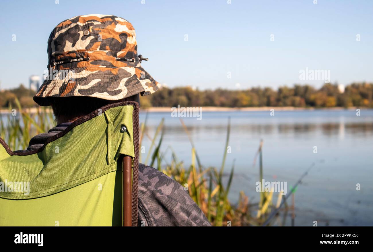 Jeune pêcheur méconnaissable attrape des poissons assis dans une chaise sur la rive d'une rivière ou d'un lac. Le pêcheur est assis sur une chaise avec du matériel de pêche. Des vacances parfaites au bord de la rivière avec une canne à pêche. Banque D'Images