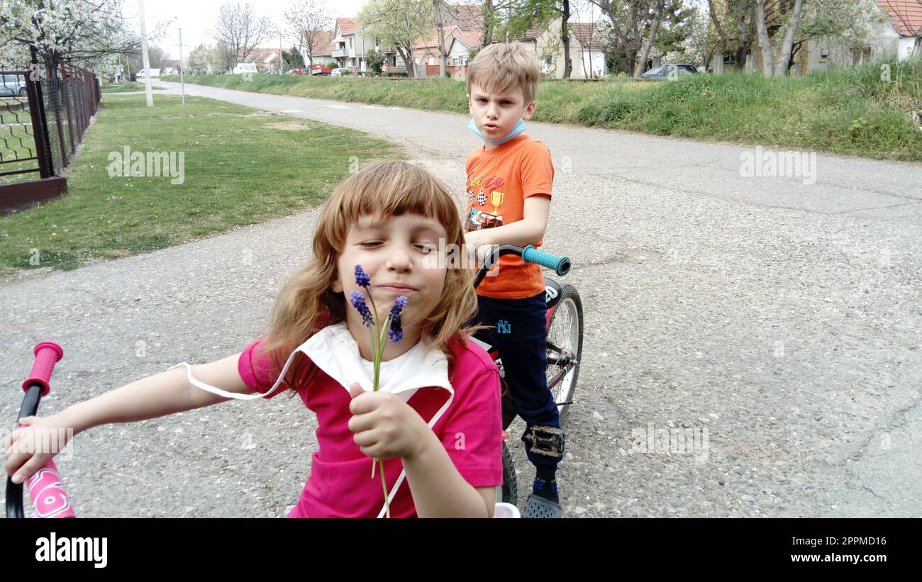 Belgrade, Serbie, le 13 avril 2020 : des enfants portant des masques chirurgicaux de protection montent à vélo. Les enfants enlevèrent leurs masques. La fille renifle une fleur violette et sourit, et le garçon fronce un sourcil et dit Banque D'Images