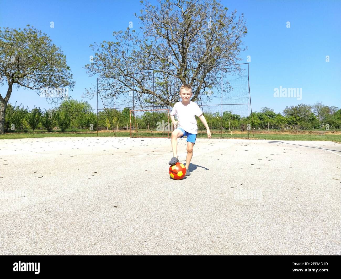Sremska Mitrovica, Serbie. 6 juin 2020. Un garçon joue au ballon sur le terrain de jeu. Terrain de sport en asphalte. Un enfant dans un t-shirt blanc. Enfant en bas âge aux cheveux blonds, 7 ans. Courir, donner des coups de pied et faire des exercices Banque D'Images