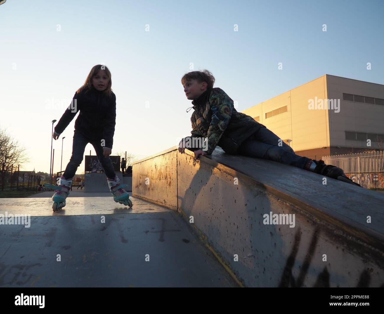Sremska Mitrovica, Serbie, 7 mars 2021. Garçon et fille sur des patins à roulettes sur le terrain de sport. Le garçon regarde la fille. La fille fait du patin à roulettes. Enfants en vêtements demi-saison. Banque D'Images
